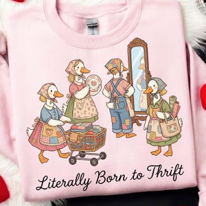 May include: Pink sweatshirt featuring a whimsical illustration of anthropomorphic geese dressed in vintage attire, engaged in thrifting activities. The phrase "Literally Born to Thrift" is printed in a cursive font below the illustration. The geese are depicted with aprons, bandanas, and carrying shopping baskets.