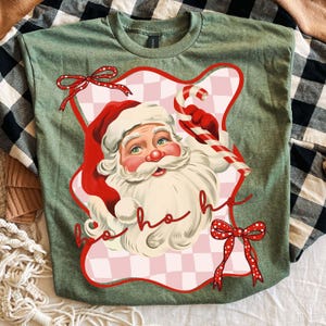 May include: Green t-shirt featuring a Santa Claus graphic. Santa is wearing a red hat and holding a candy cane. The graphic is framed by a pink and white checkered border with red bows and the words "ho ho ho".