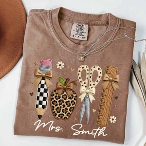 May include: A brown crew neck t-shirt featuring an embroidered design of a checkered pencil, a leopard print apple, scissors with polka dots, and a wooden ruler. Each item is accented with a tan bow. The text "Mrs. Smith" is embroidered below.
