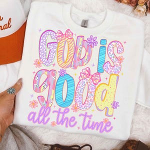 May include: White sweatshirt with the phrase "God is Good all the time" in colorful, patterned letters. The design includes bows and flowers. A trucker hat is partially visible in the upper left corner.