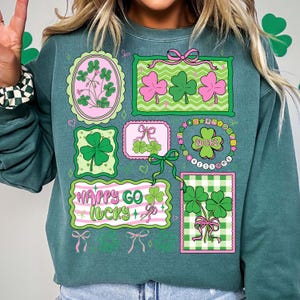 Op de afbeelding: Teal groene sweatshirt met St. Patrick's Day-ontwerpen. De afbeeldingen bevatten klavers, strikken en de tekst "Happy Go Lucky". Het ontwerp bevat roze en groene kleuren.