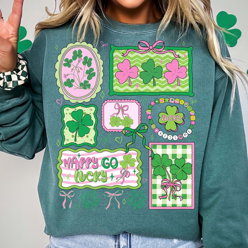 Op de afbeelding: Teal groene sweatshirt met St. Patrick's Day-ontwerpen. De afbeeldingen bevatten klavers, strikken en de tekst "Happy Go Lucky". Het ontwerp bevat roze en groene kleuren.