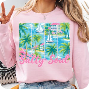 K&ouml;nnte beinhalten: Rosafarbener Pullover mit einem grafischen Druck von Palmen, Segelbooten und H&auml;usern. Der Text "Salty Soul" ist in Rosa auf der Vorderseite des Pullovers gedruckt.
