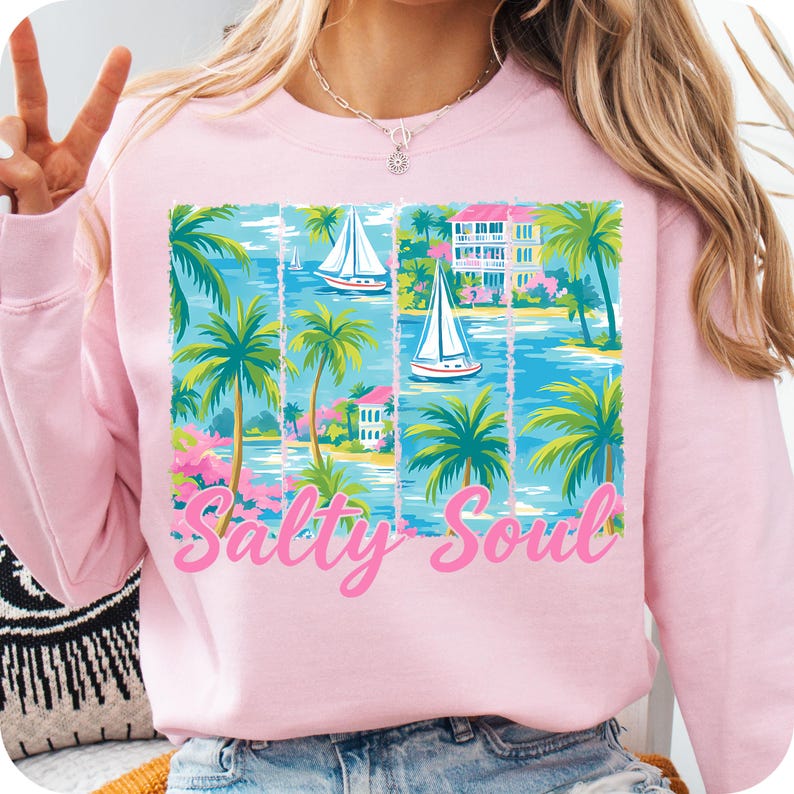 K&ouml;nnte beinhalten: Rosafarbener Pullover mit einem grafischen Druck von Palmen, Segelbooten und H&auml;usern. Der Text "Salty Soul" ist in Rosa auf der Vorderseite des Pullovers gedruckt.