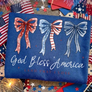 May include: A blue sweatshirt with three bow designs in red, white, and blue, with the text "God Bless America" in white script. The design is patriotic, with stars and stripes.