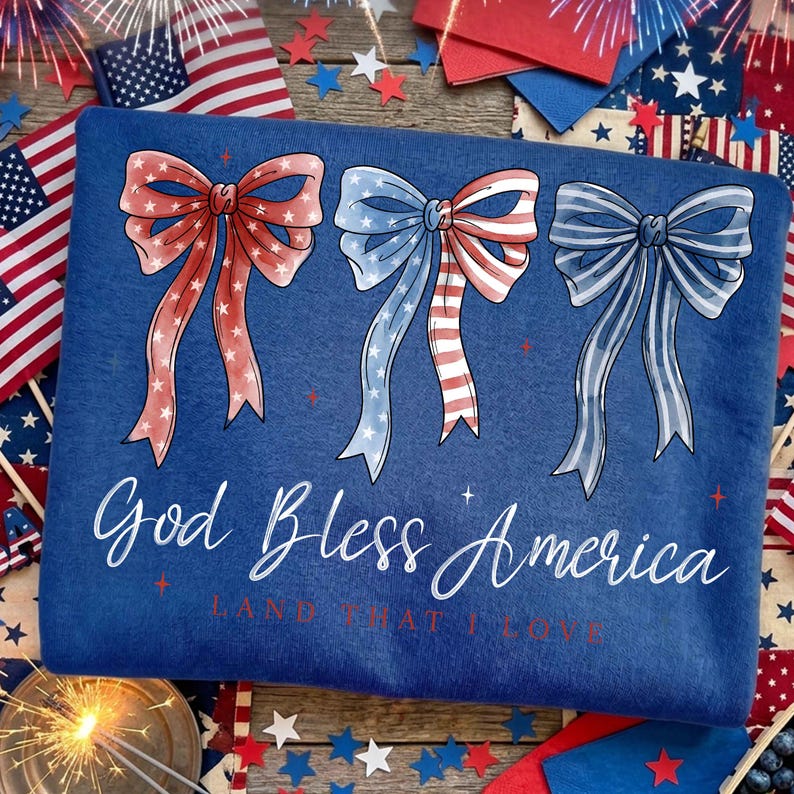 May include: A blue sweatshirt with three bow designs in red, white, and blue, with the text "God Bless America" in white script. The design is patriotic, with stars and stripes.