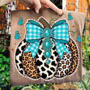 May include: A brown t-shirt featuring a pumpkin design with a turquoise and blue gingham bow, turquoise jewels, and a mix of cow and leopard print patterns. Blue butterflies and stars surround the pumpkin. The design is on a brown background.