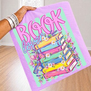 Puede incluir: Una manta de color morado claro con una pila de libros coloridos y el texto "BOOK lover" en rosa y azul. El dise&ntilde;o incluye ilustraciones de margaritas blancas, flores rosas y ramas de lavanda morada alrededor de los libros.