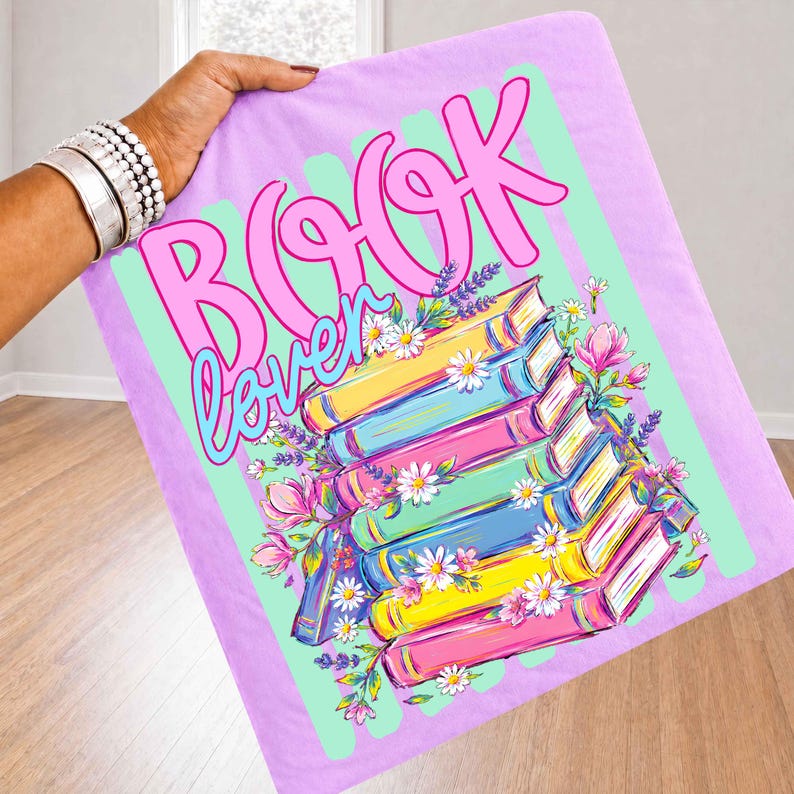 Puede incluir: Una manta de color morado claro con una pila de libros coloridos y el texto "BOOK lover" en rosa y azul. El dise&ntilde;o incluye ilustraciones de margaritas blancas, flores rosas y ramas de lavanda morada alrededor de los libros.