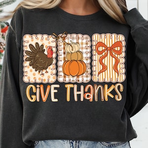 May include: Dark gray sweatshirt with a Thanksgiving-themed graphic. The graphic includes a turkey, a stack of pumpkins, and a bow, all within decorative frames. The words "GIVE THANKS" are written in orange and white letters.