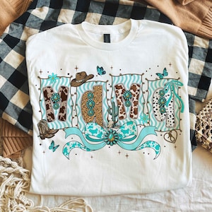 Op de afbeelding: Wit T-shirt met het woord "MAMA" in een western-thema ontwerp. De letters hebben koeienprint, turquoise accenten en cowboy-thema graphics zoals een hoed, laarzen en een cactus. Een turquoise strik staat onderaan.