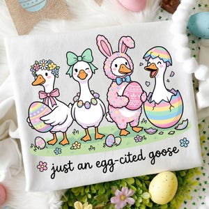 May include: White t-shirt with a whimsical Easter design. Features four cartoon geese dressed in Easter attire, holding decorated eggs, and standing on grass. The text "just an egg-cited goose" is written in a playful font.