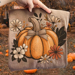 May include: A brown t-shirt featuring a large orange pumpkin with a bow, surrounded by white and orange flowers and dark green leaves. The design has a vintage, hand-drawn style, perfect for autumn.