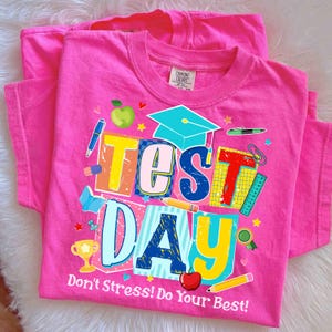 Peut inclure: Deux t-shirts rose vif avec un graphisme coloré qui dit "Test Day" avec des illustrations sur le thème de l'école. Le texte "Don't Stress! Do Your Best!" est imprimé en dessous. Les t-shirts sont en matière douce.
