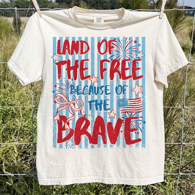 以下が含まれることがあります： クリーム色のTシャツ。愛国的なデザインで、「LAND OF THE FREE BECAUSE OF THE BRAVE」の文字が赤と青で描かれ、花火、星、アメリカ国旗が描かれています。