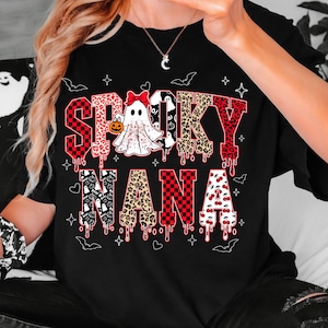May include: Black t-shirt with the words "Spooky Nana" in a mix of fonts and patterns. The word "Spooky" features red and black checkered patterns, leopard print, and dripping blood effects. A ghost with a pumpkin is in the center.