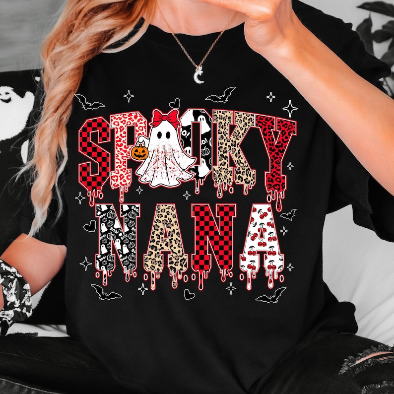 May include: Black t-shirt with the words "Spooky Nana" in a mix of fonts and patterns. The word "Spooky" features red and black checkered patterns, leopard print, and dripping blood effects. A ghost with a pumpkin is in the center.