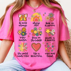 May include: Pink t-shirt with a colorful graphic design featuring flowers, butterflies, rainbows, and positive messages like "be kind always", "choose joy", "keep on blooming", "you matter", "focus on the good", "growth is a process", "the world needs you", "life is beautiful", and "you are loved".