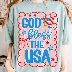 Puede incluir: Camiseta verde claro con un dise&ntilde;o patri&oacute;tico. El dise&ntilde;o presenta el texto "GOD bless THE USA" en azul, rojo y blanco, junto con una bandera estadounidense y estrellas. El dise&ntilde;o est&aacute; enmarcado por un borde rojo y rosa.