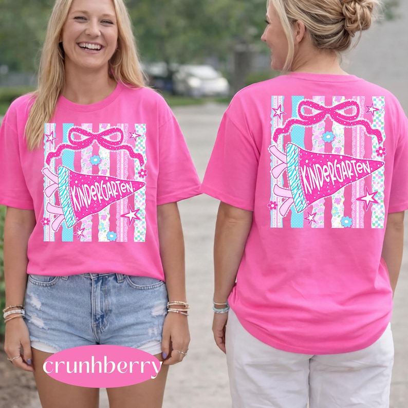 May include: A bright pink t-shirt with a graphic design featuring the word "Kindergarten" on a pennant. The design includes a bow, stars, and floral elements. The shirt is short-sleeved and has a crew neck.