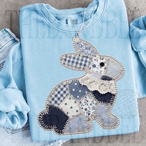 May include: A pale blue sweatshirt featuring a patchwork rabbit design. The rabbit is crafted from various blue and white patterned fabrics, including gingham and floral prints, and is edged with a pearl-like trim. The sweatshirt has the text "COMFORT COLORS" on the upper right.