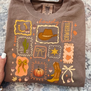 May include: A brown long-sleeved t-shirt with a western-themed graphic. The design includes a horseshoe, cowboy hat, cactus, bow, star, pumpkin, and cowboy boots. The word "howdy" is written across the top, and the word "cowgirl" is written vertically.