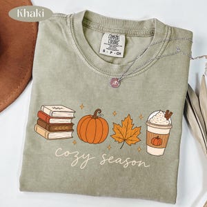 May include: A khaki crew neck t-shirt featuring a graphic print of a stack of books, an orange pumpkin, an autumn leaf, and a cup of coffee with whipped cream. The text "cozy season" is printed below the graphics in a white script font.