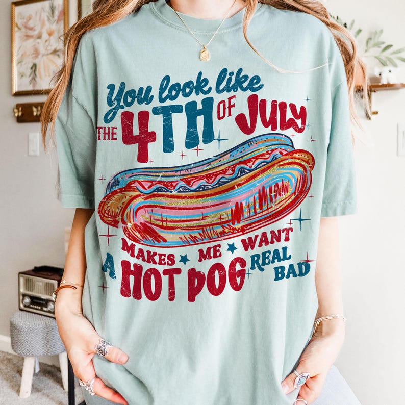 May include: A light green t-shirt with the text "You look like the 4th of July makes me want a hot dog real bad" and a colorful hot dog graphic. The design features red, blue, and pink colors.