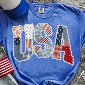 May include: A blue t-shirt with the word "USA" appliqued on the front. The letters are made of various patriotic patterned fabrics, including stars, stripes, and floral designs. A blue baseball cap is visible in the background.