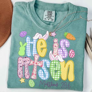 May include: A teal Comfort Colors t-shirt with the words "He is Risen" in colorful, patterned letters. Easter-themed graphics include bunny ears, eggs, and a carrot. A silver necklace with a star pendant rests on the shirt. The text "Matthew 28:6" is below.