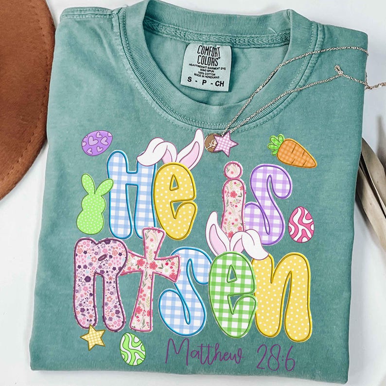 May include: A teal Comfort Colors t-shirt with the words "He is Risen" in colorful, patterned letters. Easter-themed graphics include bunny ears, eggs, and a carrot. A silver necklace with a star pendant rests on the shirt. The text "Matthew 28:6" is below.