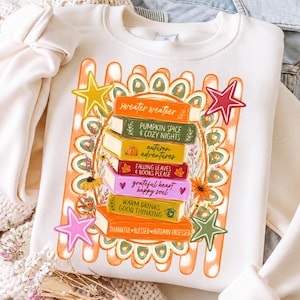 May include: A cream-coloured crewneck sweatshirt featuring a graphic print of a stack of six colourful books surrounded by floral elements and four stars. The book spines display the text: sweater weather, PUMPKIN SPICE & COZY NIGHTS, autumn adventures, FALLING LEAVES & BOOKS PLEASE, grateful heart happy soul, WARM DRINKS GOOD THINKING, and THANKFUL BLESSED AUTUMN OBSESSED.
