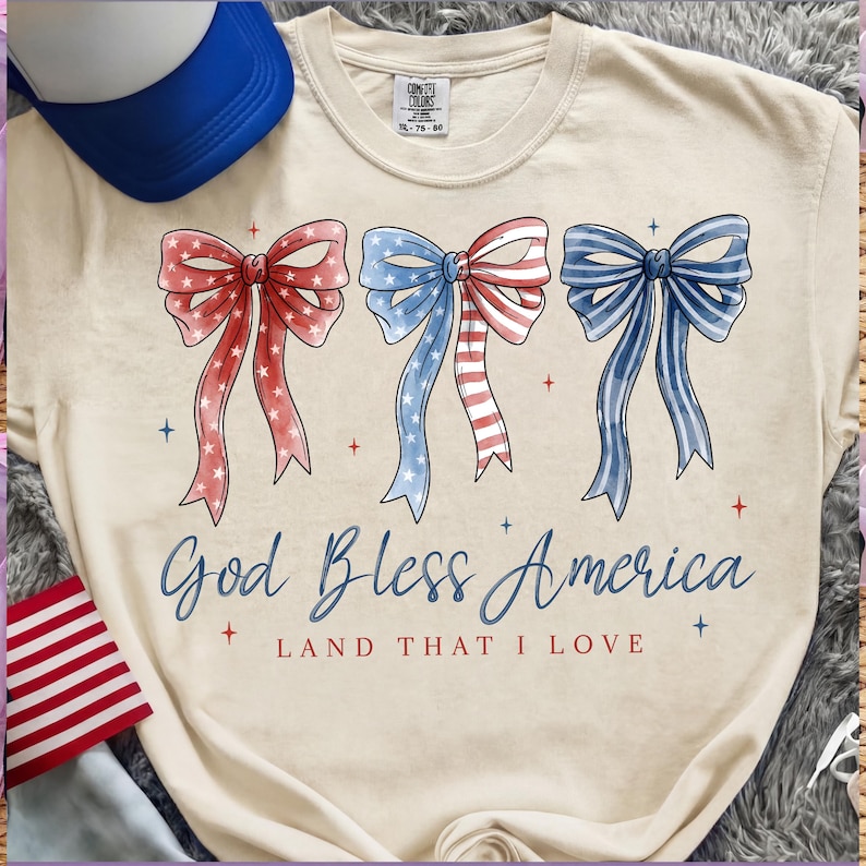 May include: Cream-colored t-shirt featuring three patriotic bow designs in red, white, and blue. The text "God Bless America" and "Land That I Love" are printed below the bows. A blue and white trucker hat is visible.