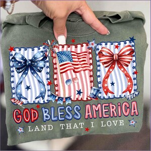 Puede incluir: Camiseta verde oliva con gr&aacute;ficos patri&oacute;ticos. El dise&ntilde;o incluye tres marcos rectangulares con un lazo azul, una bandera estadounidense y un lazo rojo. El texto "GOD BLESS AMERICA LAND THAT I LOVE" est&aacute; impreso en rojo y azul.