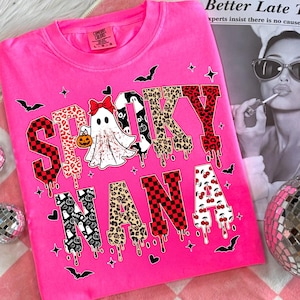 May include: A hot pink sweatshirt with the words "Spooky Nana" in a mix of patterns and fonts. The letter designs include leopard print, cherries, and checkered patterns. A ghost with a pumpkin is in the center. Bats and stars are scattered around the text.