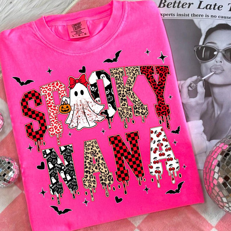 May include: A hot pink sweatshirt with the words "Spooky Nana" in a mix of patterns and fonts. The letter designs include leopard print, cherries, and checkered patterns. A ghost with a pumpkin is in the center. Bats and stars are scattered around the text.