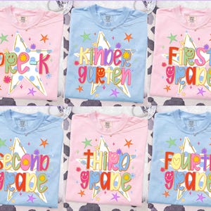May include: A collection of six t-shirts in light pink and light blue, each featuring a large white star with blue polka dots. The shirts display colorful text for school grades including Pre-K, Kindergarten, First, Second, Third, and Fourth Grade.