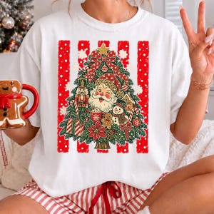 Può includere: Una t-shirt bianca a maniche corte con la grafica di un albero di Natale decorato con il volto di Babbo Natale, uno schiaccianoci, un pupazzo di neve, un omino di pan di zenzero, regali e un grande fiocco rosso su sfondo a righe bianche e rosse.