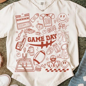 May include: Cream-colored t-shirt with a red graphic design featuring football-related icons and the words "GAME DAY." The design includes a football, helmet, trophy, and a football field.