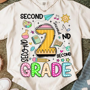 Pu&ograve; includere: T-shirt color crema con un design grafico colorato per la seconda elementare. Il design include un grande numero 2 a forma di matita gialla, le parole "SECOND GRADE" e varie illustrazioni scolastiche.