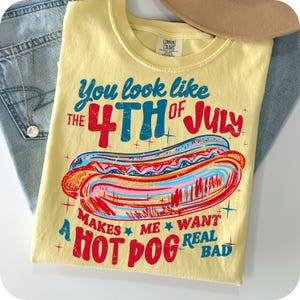 May include: Yellow t-shirt with the text "You look like the 4th of July" and a graphic of a hot dog. The shirt also says "Makes me want a hot dog real bad". The shirt is folded on top of blue jeans.