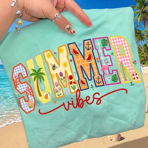 Puede incluir: Una camiseta de color verde azulado claro con las palabras "SUMMER vibes" en una fuente decorativa y colorida. Las letras están llenas de imágenes temáticas de verano como una palmera, rodajas de sandía y un flamenco. La camiseta es sostenida por una mano.
