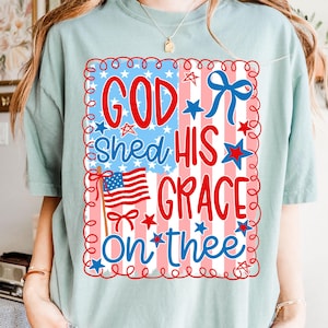May include: A light blue t-shirt with a patriotic design. The graphic features the text "GOD Shed HIS GRACE On *thee" in red and blue, along with an American flag and stars. The design is framed with a red border.