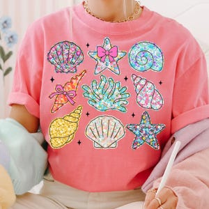 May include: A pink t-shirt with a floral pattern featuring seashells, starfish, and coral. The design is colorful and whimsical.