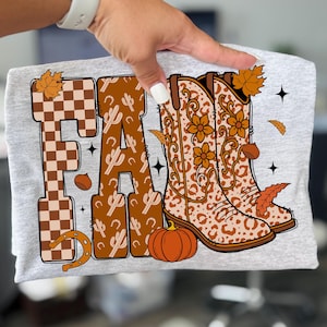 May include: A light grey t-shirt featuring the word "FALL" in brown and tan. The "F" is checkered, the "A" has a cactus pattern, and the "L" has cowboy boots, a pumpkin, and autumn leaves.