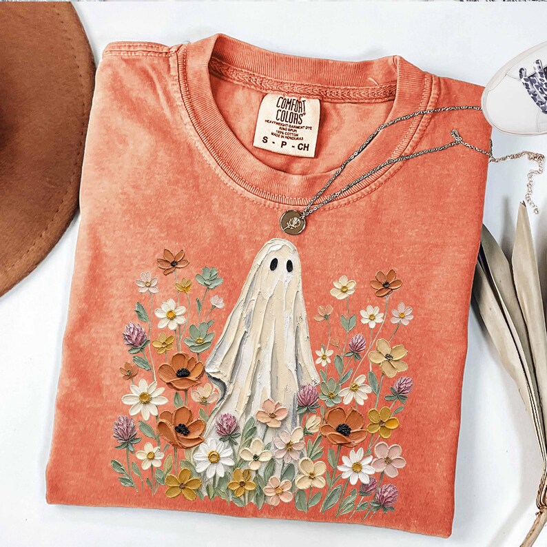 May include: A coral-colored t-shirt featuring a central graphic of a white ghost surrounded by a variety of colorful painted-style flowers. The shirt has a crew neckline with a Comfort Colors label and a thin silver chain necklace resting on it.