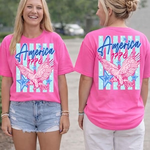 Puede incluir: Una camiseta rosa brillante con un dise&ntilde;o patri&oacute;tico. La parte delantera y trasera presentan la palabra "America" sobre "1776" y un &aacute;guila rosa. El dise&ntilde;o incluye estrellas y rayas azules.