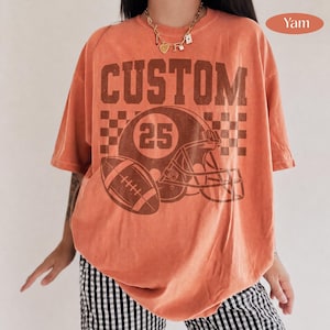 May include: An orange short-sleeve t-shirt featuring a dark brown graphic of a football helmet with the number 25, a football, and a checkered pattern. The word CUSTOM is printed in large dark brown block letters above the helmet graphic.