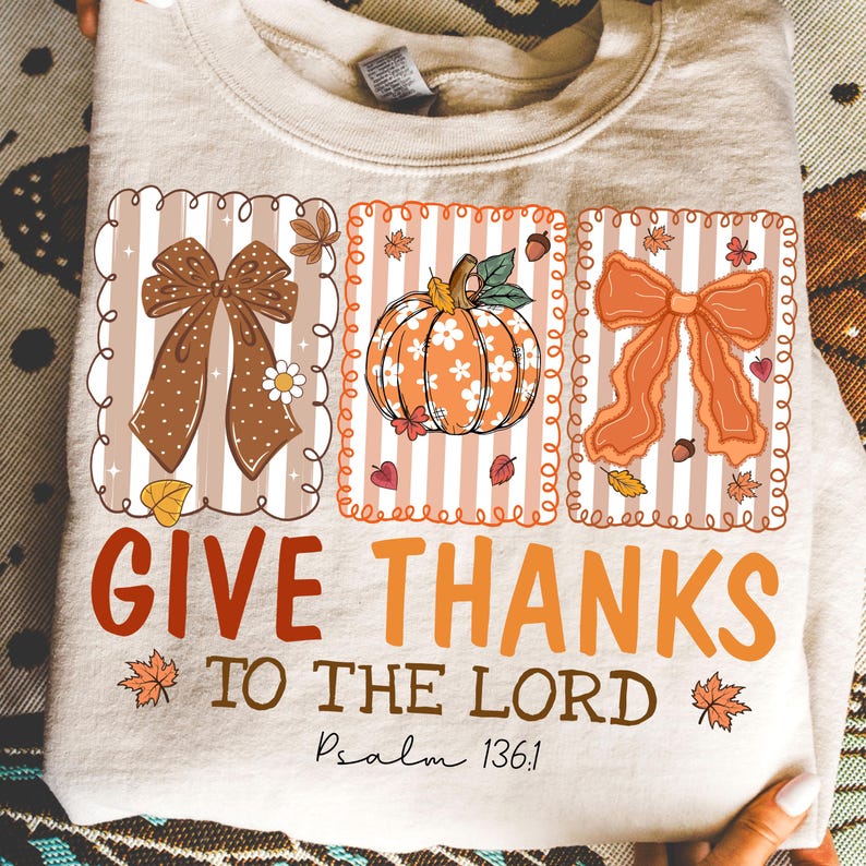 May include: A beige sweatshirt with a fall-themed graphic. The design includes three framed images of a bow, a pumpkin, and another bow. The words "GIVE THANKS TO THE LORD" are printed in orange and brown, with "Psalm 136:1" below.