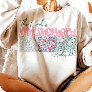 May include: A white sweatshirt with the text "The Lord is My Shepherd" in pink lettering and a floral pattern below. The text "Psalm 23:1" is also included.